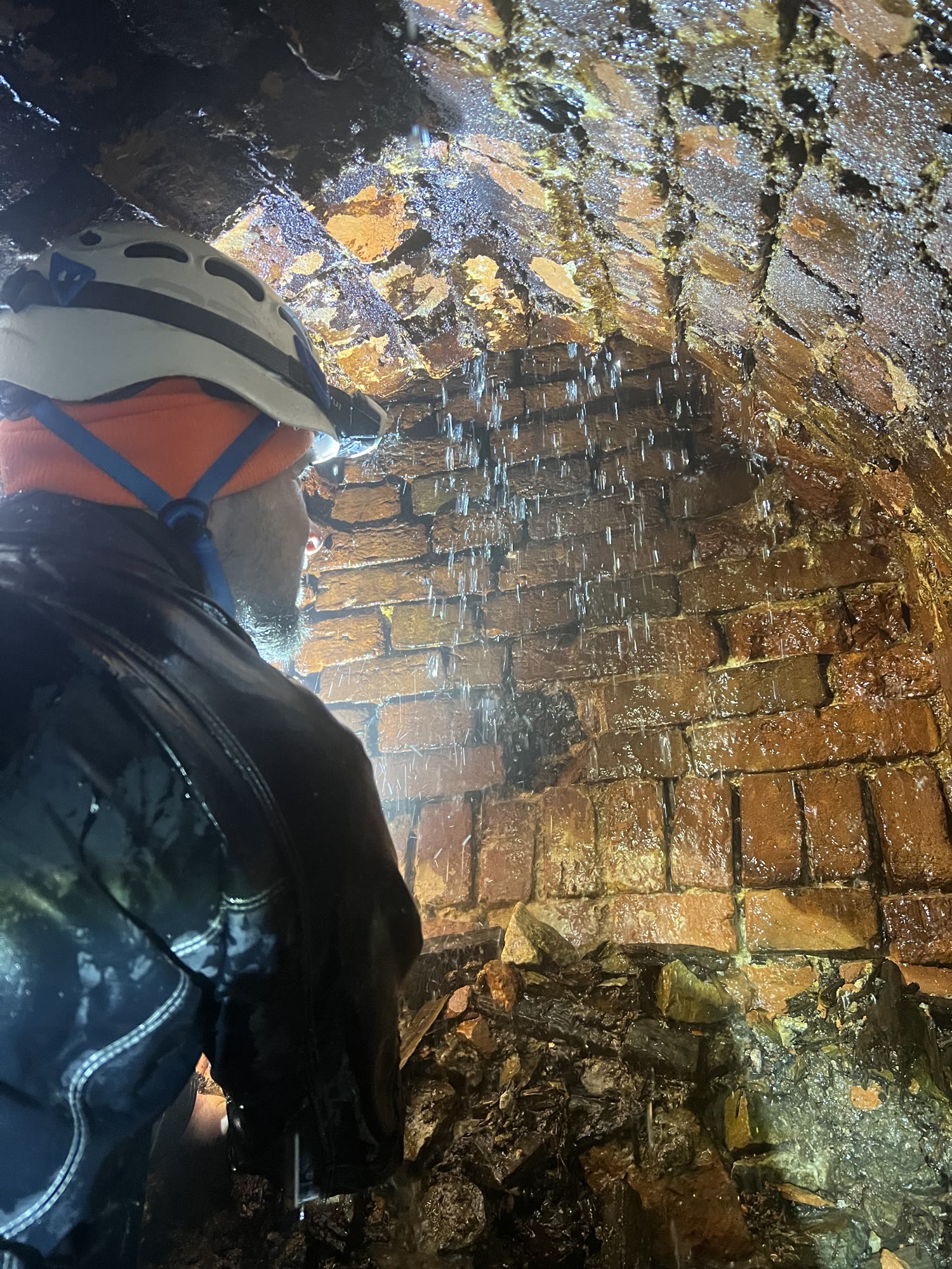 A caver in a helmet looks up at the brick vault at the end of the drained tunnel, water dripping from the ceiling